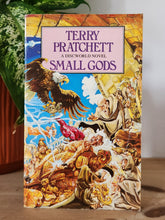 Load image into Gallery viewer, Book titled 'Small Gods' by Terry Pratchett on a wooden surface with a plant in the background
