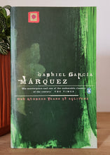 Load image into Gallery viewer, Book titled 'One Hundred Years of Solitude' by Gabriel Garcia Marquez on a wooden surface with a plant in the background.