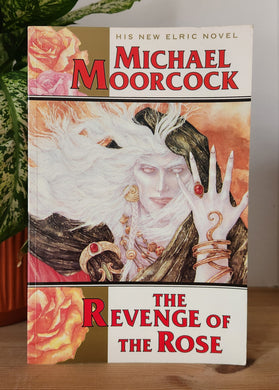 Book titled 'The Revenge of the Rose' by Michael Moorcock on a wooden surface with a plant in the background.