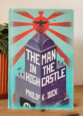 Book titled 'The Man in the High Castle' by Philip K. Dick on a wooden surface with a plant in the background.