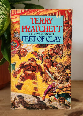Book titled 'Feet of Clay' by Terry Pratchett on a wooden surface with a plant in the background